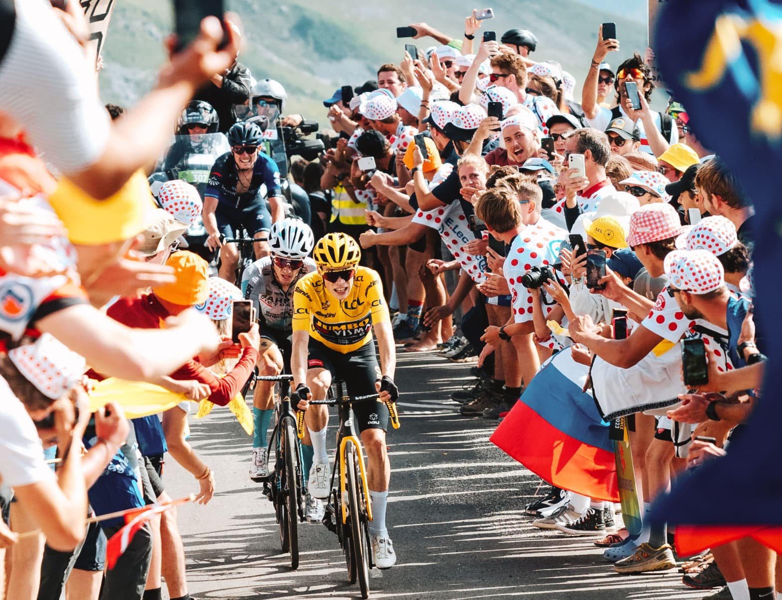 Jonas Vingegaard, followed by members of the peloton pedals his way up a narrow road. He is surrounded by a mass of excited fans in polka dot shirts and hats waving signs and flags.